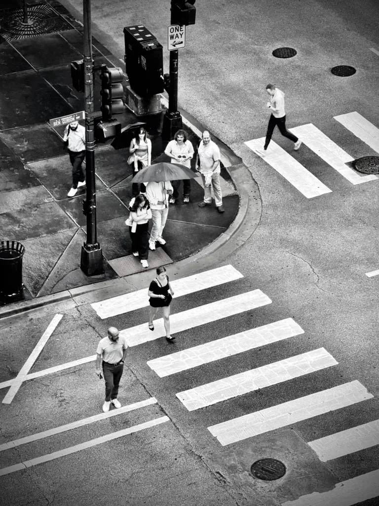 Black and white view from above of people crossing a wet crosswalk, one holding an umbrella