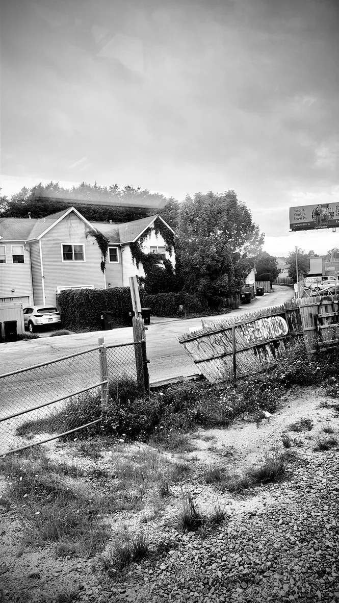 Black and white view of houses, a leaning fence and a gravel lot under a cloudy sky