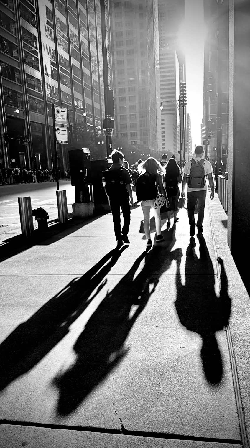 Black and white street scene of people walking into the sun, their long shadows stretching across the sidewalk